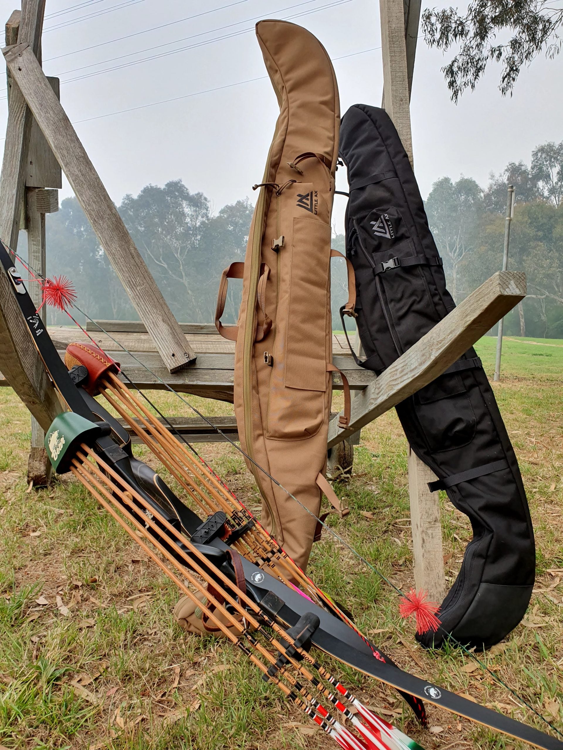 Two Little Ax bow cases in coyote brown and black standing against a weathered timber fence rail in misty morning light, gum trees and paddock behind, with a black recurve bow, green quiver and wooden arrows in the foreground